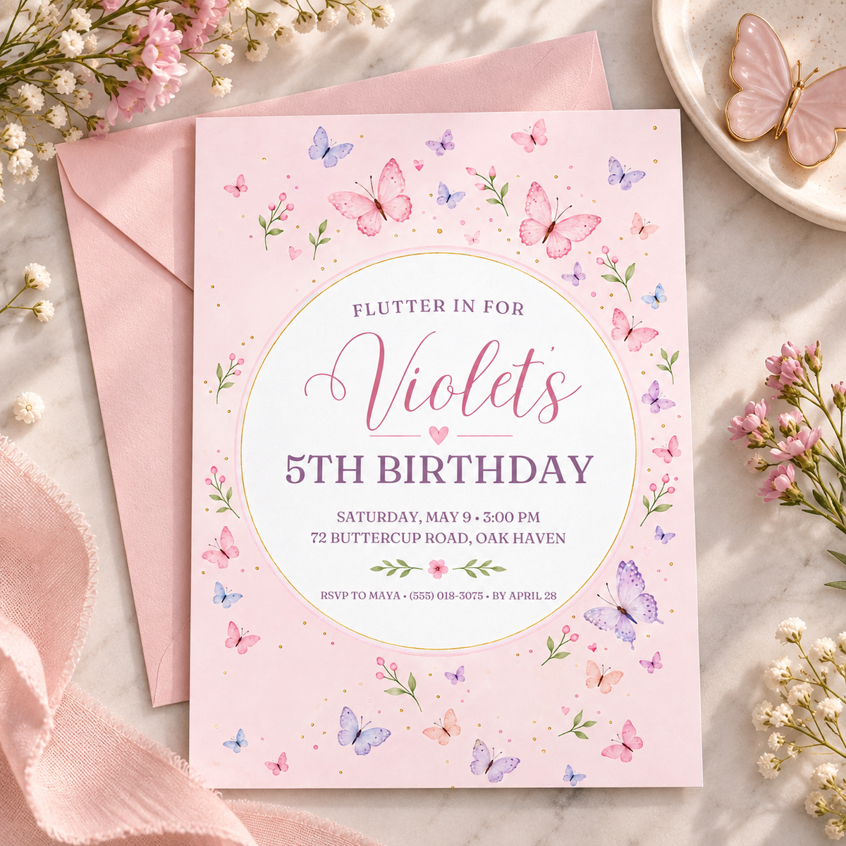 Blush pink butterfly invitation beside a matching pink envelope and flowers