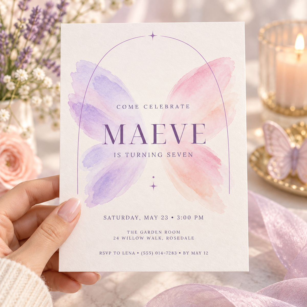 Hand holding a white invitation with soft lilac and pink watercolor butterfly wings behind the name