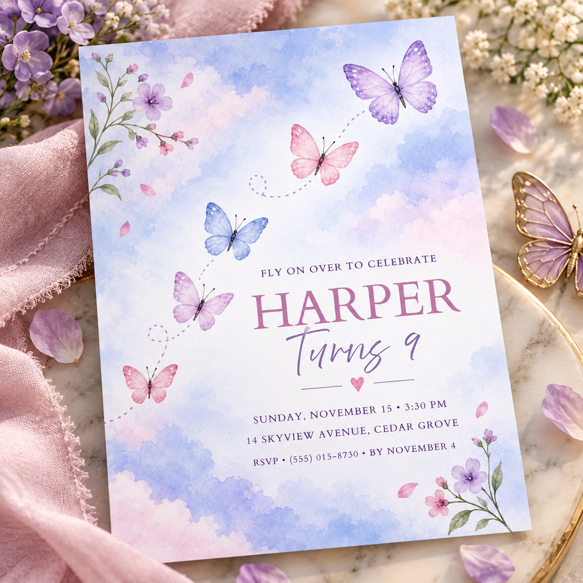 Pastel watercolor butterfly invitation angled on pink linen beside a gold butterfly
