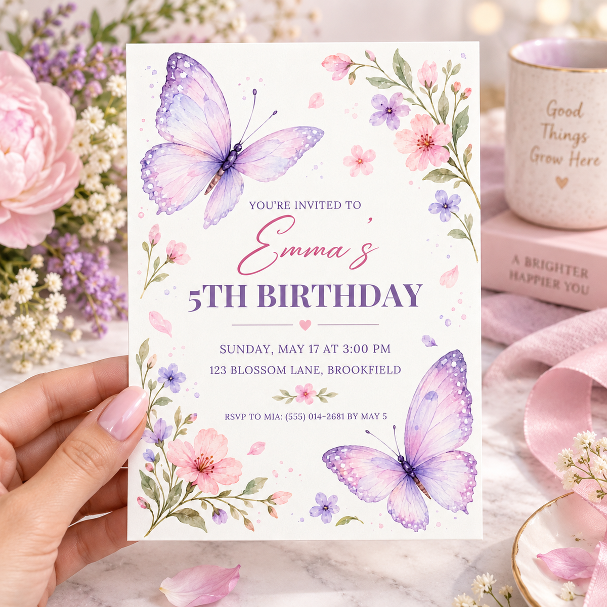 Hand holding a purple butterfly birthday invitation with pink blossom sprigs and sample details
